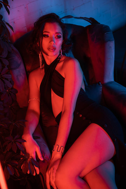Sexy and trendy asian woman sitting in armchair near plants in colorful neon light in night club - Photo, Image