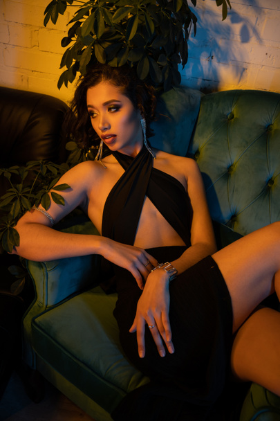 Fashionable asian woman in evening dress sitting on couch near plants in night club with lighting - Photo, image