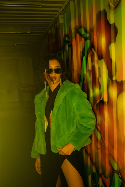 Bella donna asiatica in occhiali da sole e giacca di pelliccia sintetica in piedi vicino graffiti in night club - Foto, immagini