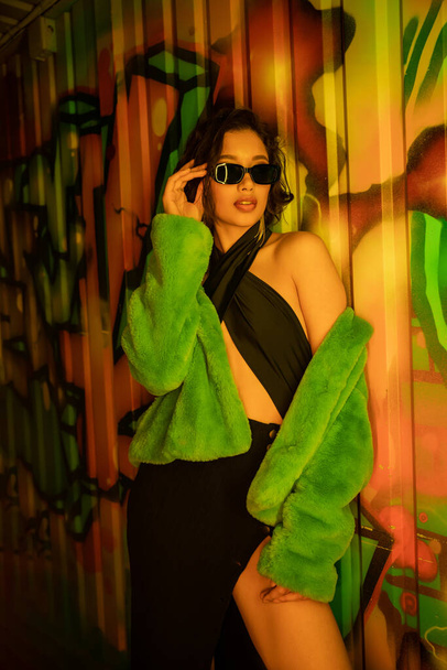 Sexy asian woman in fake fur jacket touching sunglasses and standing near graffiti in night club - Foto, Imagem