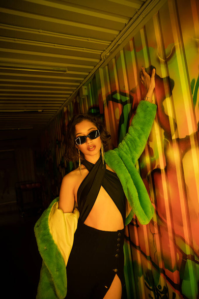 Sexy young asian woman in sunglasses and dress posing near graffiti on wall in night club - Photo, Image