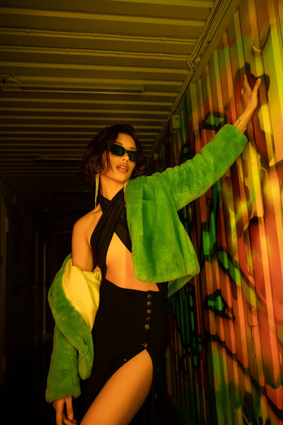 Pretty and sexy asian woman in sunglasses and faux fur jacket posing near graffiti in night club - Фото, зображення