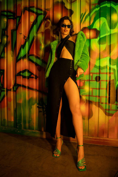 Trendy asian woman in heels, dress and sunglasses standing near graffiti on wall in night club - Foto, imagen