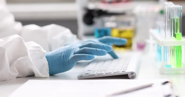 Wetenschapper chemicus in beschermend pak en handschoenen typen op toetsenbord in de buurt van reageerbuizen in laboratorium 4k film. Ontwikkeling van het concept "vaccin tegen covid19" - Video