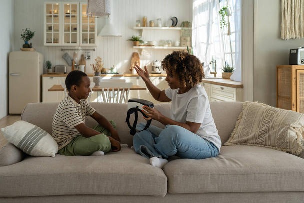 Joven madre afroamericana sosteniendo casco VR introduciendo la tecnología de realidad virtual a su hijo mientras pasan tiempo libre juntos en casa. Mamá padre explorando mundo virtual con niño - Foto, Imagen