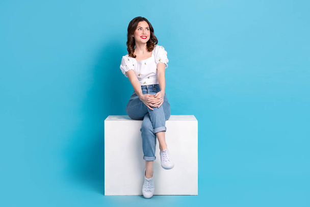 Full length photo of dreamy shiny girl dressed white top sitting cube looking empty space isolated blue color background. - Photo, image