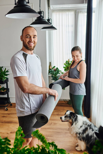 Happy man in sportswear looking at camera near girlfriend with fitness mat and border collie at home - 写真・画像