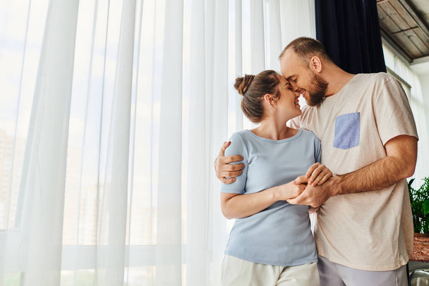 Side view of smiling couple in homewear holding hands and hugging while standing at home - Photo, Image