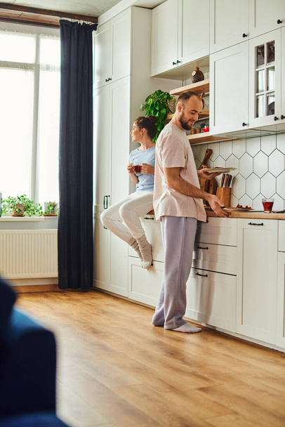 自宅でキッチンでお茶を保持しているガールフレンドの近くでおいしい朝食でプレートを取るホームウェアの男 - 写真・画像