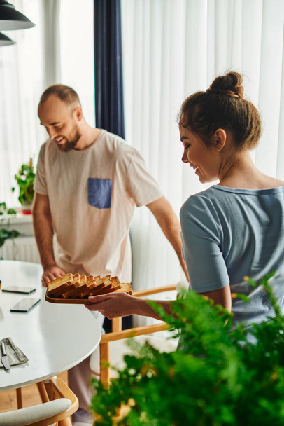Smiling woman in homewear holding toasts near blurred boyfriend during breakfast at home in morning - Фото, зображення