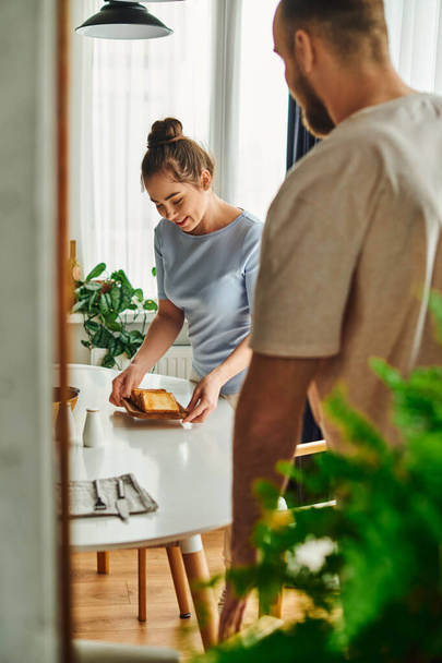 glimlachende vrouw in huiskleding zetten van toast op tafel in de buurt wazig vriendje thuis in de ochtend - Foto, afbeelding