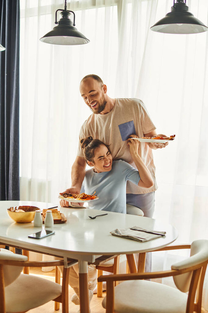 Positive woman in homewear sitting near boyfriend putting breakfast on table at home in morning - Photo, Image