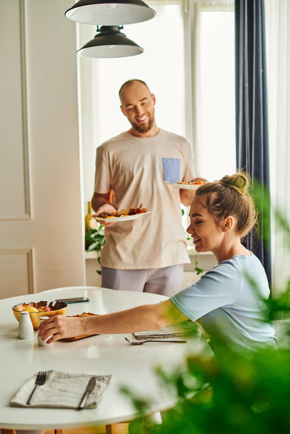 Smiling woman taking salt near blurred boyfriend with breakfast on plates at home in morning - Fotografie, Obrázek