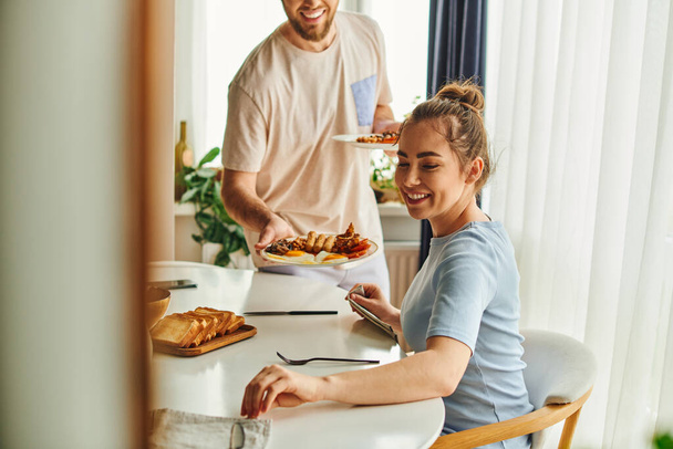 Sorrindo mulher sentada perto de talheres e namorado borrado segurando saboroso café da manhã em casa de manhã - Foto, Imagem