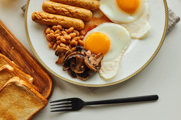 自宅でテーブル上のカッティングボード上のトーストの近くの揚げ卵とソーセージとの朝食のトップビュー - 写真・画像