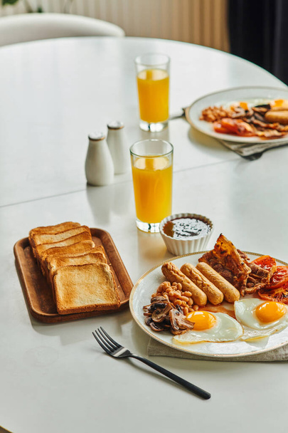 Традиционный завтрак с яичницей, сосисками и беконом рядом с апельсиновым соком и джемом на столе дома - Фото, изображение