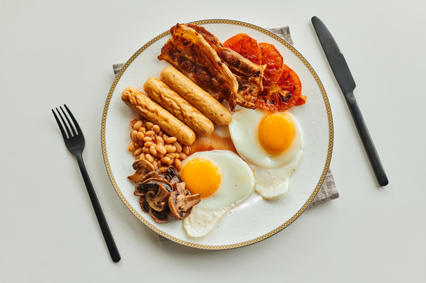 Вид на утреннюю трапезу с сосисками, жареными яйцами и беконом на тарелке возле столовых приборов на столе - Фото, изображение