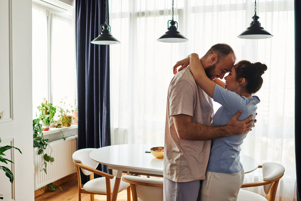 Πλευρική άποψη του χαμογελαστού ζευγαριού σε homewear αγκαλιάζει ο ένας τον άλλον και να περνούν χρόνο στο σπίτι - Φωτογραφία, εικόνα