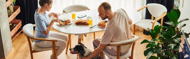 Hombre sonriente en ropa de casa acariciando frontera collie mientras desayuna con su novia en casa, pancarta - Foto, imagen