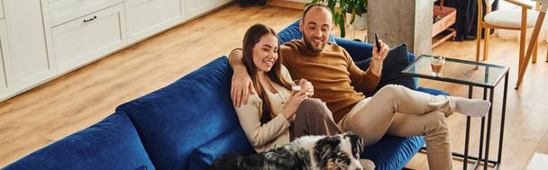 Smiling couple with coffee and remote controller sitting near border collie on couch at home,banner - Foto, Bild