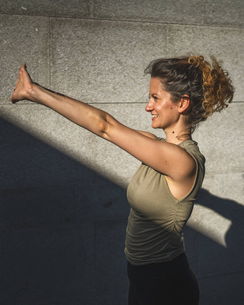 Uma mulher caucasiana adulta fazendo ioga prática esticar no dia de verão ao ar livre na cidade em frente à parede com sombra e sol estilo de vida saudável conceito de bem-estar auto cuidado - Foto, Imagem