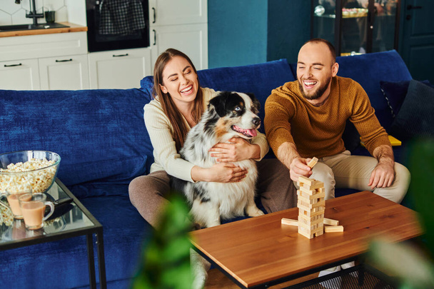Positives Paar verbringt Zeit mit Holzklötzchen-Spiel und Border Collie auf der Couch neben Popcorn zu Hause - Foto, Bild