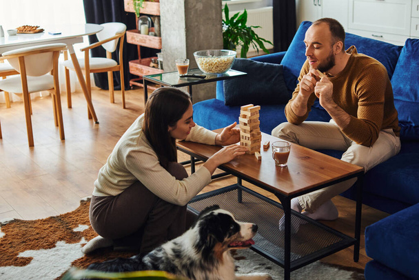 Smiling man spending time with girlfriend playing wood blocks game near border collie at home - Fotó, kép