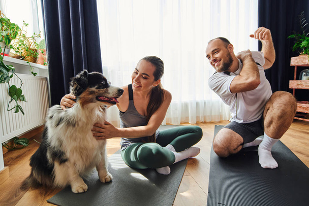Smiling man warming up near girlfriend in sportswear petting border collie on fitness mat at home - Foto, Imagem