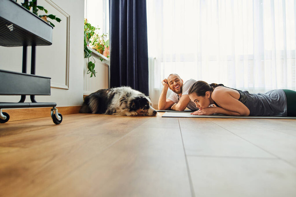 Uomo sorridente in abbigliamento sportivo guardando fidanzata e confine collie mentre sdraiato sul tappeto fitness a casa - Foto, immagini