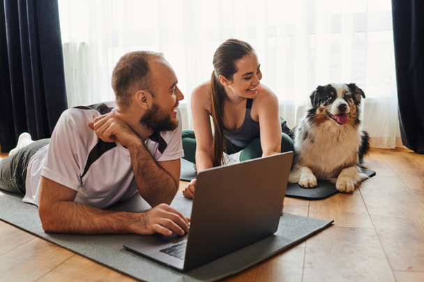 Positive couple in sportswear using laptop on fitness mats and looking at border collie at home - Photo, Image