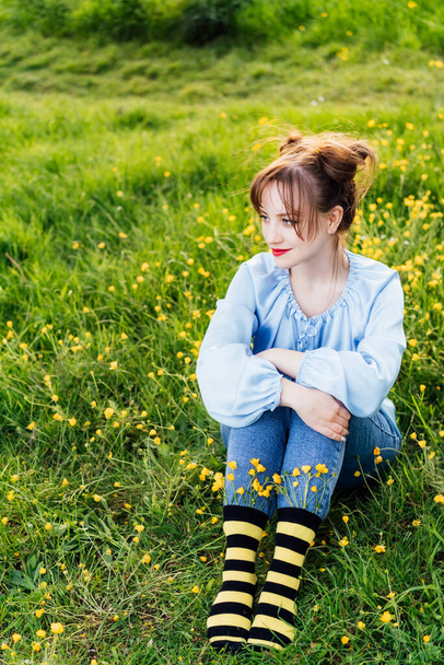 Νεαρή γυναίκα που φοράει μπλε μπλούζα, τζιν και ριγέ μαύρες και κίτρινες κάλτσες με λουλούδια μέσα κάθεται στο πράσινο γρασίδι του ανθισμένου λιβαδιού. Στυλ Anime. Έννοια της προστασίας των μελισσών, εποχή άνθισης. - Φωτογραφία, εικόνα