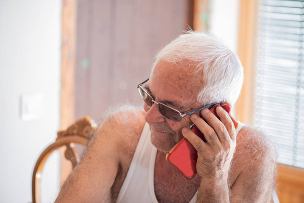 мужчина в возрасте восьмидесяти лет в белом майке разговаривает на бордовом мобильном телефоне, седые волосы и коричневая кожа, улыбаясь. - Фото, изображение
