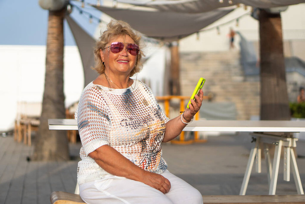У сонячний день біля пляжу, приваблива літня леді з білим волоссям посміхнулася, коли вона читала повідомлення на своєму смартфоні. Старша жінка насолоджувалася вільним часом і відпусткою, тримаючи мобільний телефон - Фото, зображення