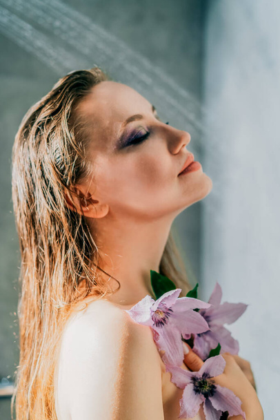 hermosa hembra desnuda delgada sexy con el pelo largo y la piel sana en la ducha. Mujer seductora con los ojos cerrados maquillaje púrpura y cuerpo desnudo perfecto cubierto de flores púrpura disfruta del momento - Foto, imagen