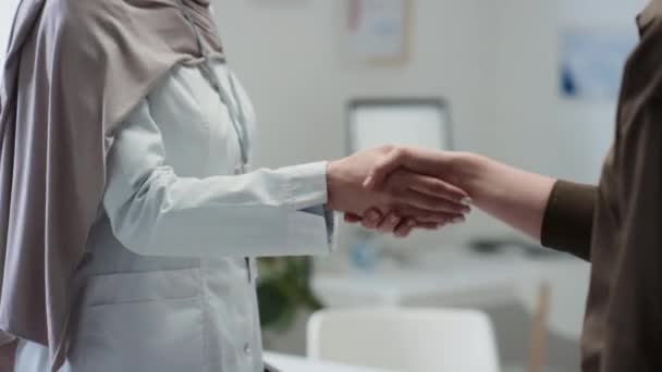 Крупный план молодой женщины-врача в хиджабе и лабораторном халате, пожимающей руку врачу на рабочем месте, поздравляя друг друга с хорошими результатами лечения и уходом из медицинского кабинета - Кадры, видео