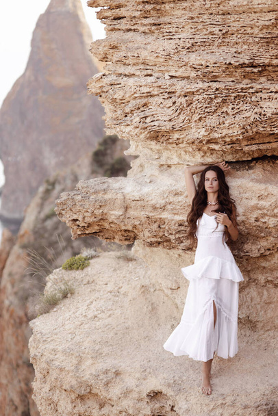 μόδα ταξίδια φωτογραφία της όμορφης γυναίκας με σκούρα μαλλιά σε κομψό λευκό φόρεμα ποζάρουν σε εκπληκτικό τόπο της φύσης, τοπίο με βουνά και τη θάλασσα - Φωτογραφία, εικόνα