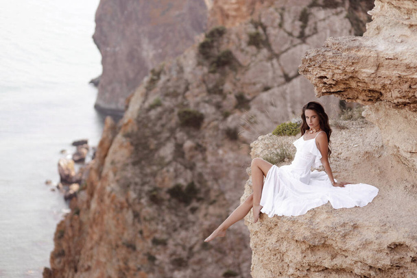 Modereise Foto von schönen Frau mit dunklen Haaren in elegantem weißen Kleid posiert in erstaunlicher Natur Ort, Landschaft mit Bergen und Meer - Foto, Bild