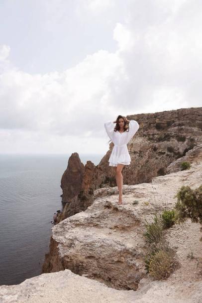 fashion travel photo of beautiful woman with dark hair in elegant white dress posing in amazing nature place, landscape with mountains and sea - Foto, afbeelding