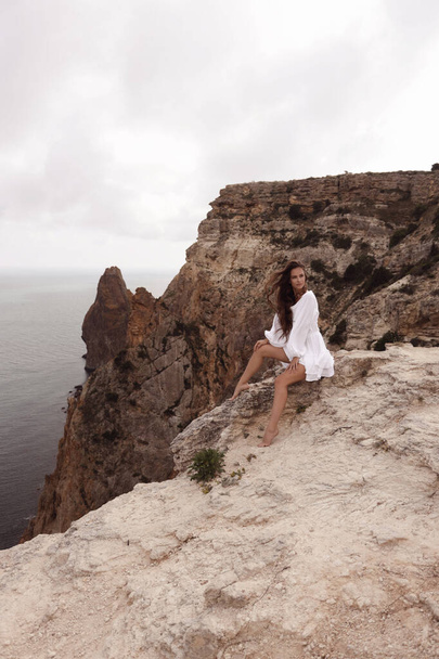 fashion travel photo of beautiful woman with dark hair in elegant white dress posing in amazing nature place, landscape with mountains and sea - Photo, image