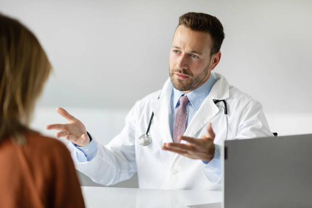 Vakava mies GP puhuu naispotilaan kanssa kokouksen aikana klinikalla, mies lääkäri valkoisessa univormussa keskustella lääkärintarkastuksen tuloksista naisen kanssa - Valokuva, kuva