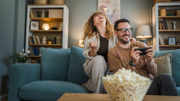 Adult couple man and woman caucasian husband and wife or boyfriend and girlfriend he play video games at home hold mobile phone smart phone have fun leisure joy and bonding concept copy space - Photo, Image