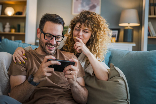Dorosłe para mężczyzna i kobieta kaukaski mąż i żona lub chłopak i dziewczyna on grać w gry wideo w domu hold telefon komórkowy smartfon zabawy rekreacji radość i więzi koncepcja kopiowanie przestrzeni - Zdjęcie, obraz