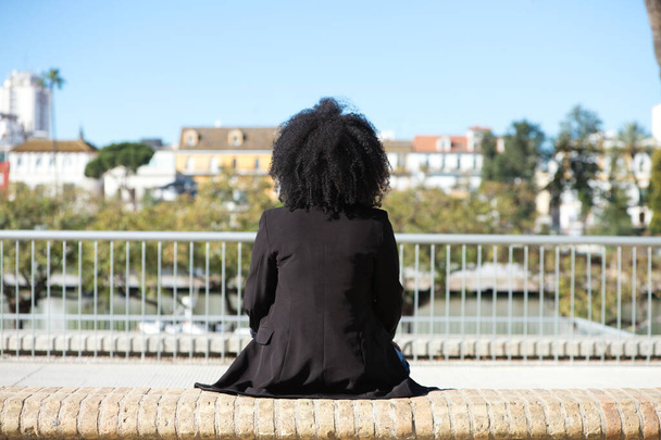 joven, hermosa mujer negra con el pelo afro sentado en un banco mirando hacia el otro lado. Foto tomada por detrás. - Foto, imagen