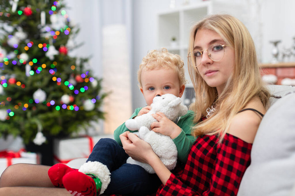 The woman is wearing an elegant plaid dress. The woman is holding a small child on her lap. Both are sitting on a gray couch. The room is decorated with Christmas decorations. The woman is wearing - Foto, imagen