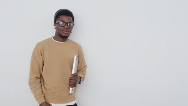 Porträt eines selbstbewussten jungen Afroamerikaners in stilvoller Freizeitkleidung mit Mütze und Brille, Laptop in die Kamera haltend, weißer Wandhintergrund - Filmmaterial, Video