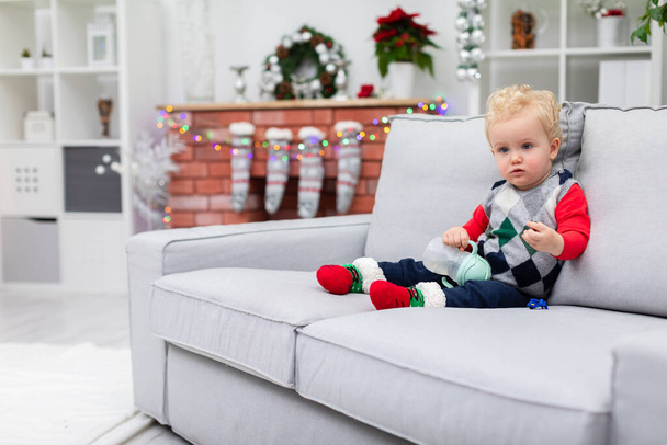The boy is sitting on a gray couch. In the background you can see a brick fireplace with Christmas decorations hanging on it: colorful lights and Christmas socks. The child is wearing a colorful - Fotoğraf, Görsel