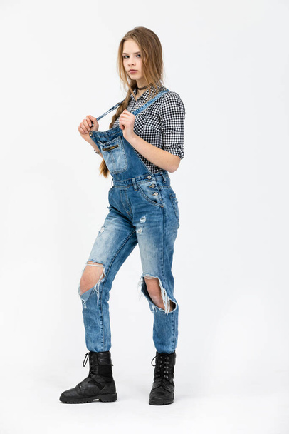 A young girl poses for a photo in a standing position., The girl is wearing a checked shirt and rubbed jeans. The girl is wearing black high top shoes. She is styled in a braid and has disheveled hair - Фото, изображение