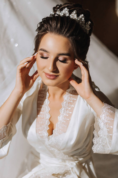 The brunette bride, dressed in a satin robe, sits on the couch. Beautiful hair and makeup. Wedding portrait. Sincere smile. A beautiful bride in a dressing gown is posing - Fotografie, Obrázek
