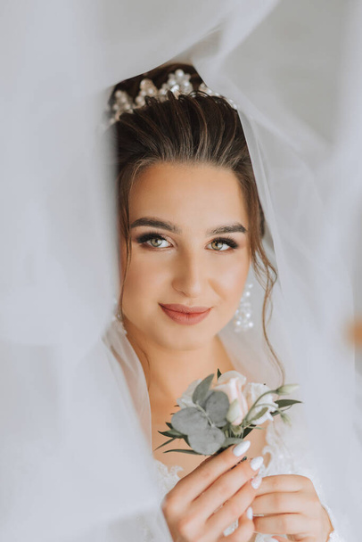 A brunette bride in a white dress holds her wedding bouquet, poses. French manicure. Open shoulders. Beautiful hands. Long veil. Morning of the bride. Details - Photo, Image