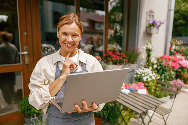 Belle propriétaire de magasin de fleurs portant tablier travaillant sur ordinateur portable dans son magasin - Photo, image